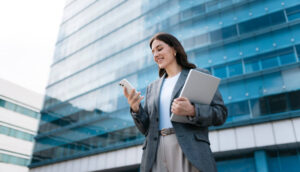Pessoa vestindo blazer cinza e calça clara, segurando um notebook prateado em um braço e um smartphone na outra mão, em frente a um prédio moderno com fachada de vidro azul