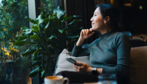 Pessoa sentada em um sofá próximo a uma janela com plantas, segurando um smartphone na mão direita