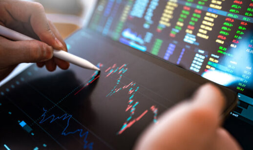 Close de mãos utilizando caneta digital para analisar gráficos de ações em um tablet, com tela ao fundo exibindo cotações e indicadores do mercado financeiro