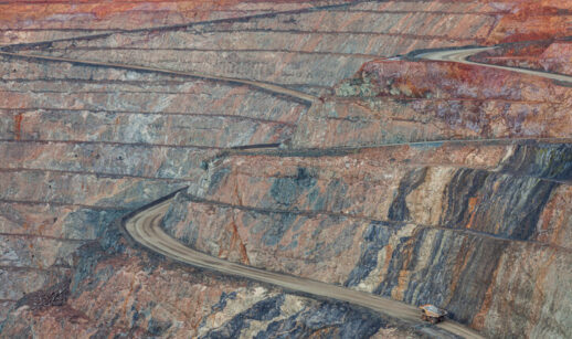 Vista de uma mina a céu aberto com estradas em curvas ao longo das paredes escalonadas, mostrando diferentes camadas de rochas em tons de marrom, cinza e vermelho