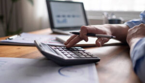 Mão segurando uma caneta enquanto digita em uma calculadora sobre uma mesa com documentos financeiros