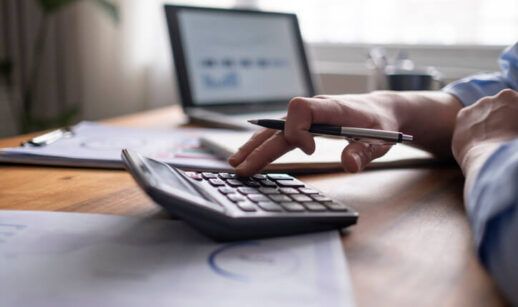Mão segurando uma caneta enquanto digita em uma calculadora sobre uma mesa com documentos financeiros